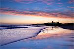 Looking across Embleton Bay at sunrise towards the silhouetted ruins of Dunstanburgh Castle in the distance and the vivid colours in the sky reflecting in the sea and wet sand, Embleton, near Alnwick, Northumberland, England, United Kingdom