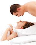 Young man looking down on young smiling woman laying in bed, covered with white quilt