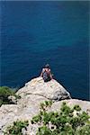 Lonely young hiker sitting on top of the rock and looking to the blue sea