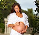 Pregnant woman on a white clothes standing on the middle of the trees touching her belly