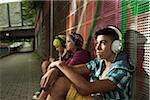 Children sitting next to wall outdoors, wearing headphones and listening to music, Germany