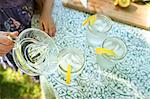 Making Lemonade. Overhead Shot Of Lemonade Glasses With A Fresh Slice Of Lemon In The Edge Of The Glass. A Child Pouring The Drink From A Jug.