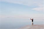 A Man Standing At Edge Of The Flooded Bonneville Salt Flats At Dusk, Taking A Photograph With A Tablet Device, Near Wendover.