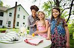 A Summer Family Gathering At A Farm. A Woman And Two Children Standing Outside By A Table, Laying The Table. Making Lemonade.