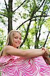 A Young Girl In A Pink Patterned Sundress Sitting On The Grass Under The Trees In A Farmhouse Garden.