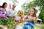 Family Party. A Table Laid With Salads And Fresh Fruits And Vegetables. Parents And Children. A Child Holding Out Fresh Picked Carrots.