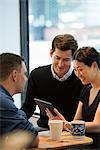 A Group Of People Sitting Around A Table In A Coffee Shop. Looking At The Screen Of A Digital Tablet.