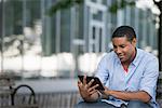 Summer In The City. People Outdoors, Keeping In Touch While On The Move. A Man Sitting On A Bench Using A Digital Tablet.