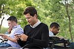 Summer In The City. One Woman And Three Men Sitting In The Park, Each On Their Own Phone Or Using A Tablet.