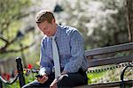 A Businessman In A Shirt With White Tie, Sitting On A Park Bench Under The Shade Of A Tree With Blossom.