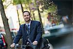 A Man In A Business Suit, Outdoors In A Park. Sitting On A Bicycle.