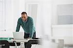 Business. A Man Standing Over A Desk, Leaning Down To Use A Laptop Computer.