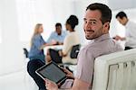 Office Interior. Meeting. One Person Looking Over His Shoulder And Away From The Group. Holding A Digital Tablet.