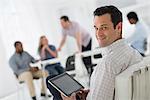 Office Interior. Meeting. One Person Looking Over Her Shoulder And Away From The Group. Holding A Digital Tablet.