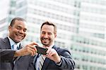 City. Two Men In Business Suits, Looking At A Smart Phone, Smiling.