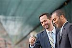 City. Two Men In Business Suits, Looking At A Smart Phone, Smiling.