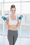 Happy athletic blonde exercising with dumbbells in bright room