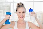 Smiling sporty blonde holding flask and dumbbell in bright room