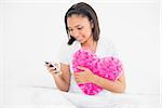 Pleased dark haired model cuddling a pillow and using a mobile phone in bright bedroom