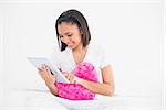 Concentrated young dark haired model holding a pillow and using a tablet pc in bright bedroom