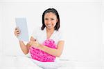 Smiling young dark haired model cuddling a pillow and using a tablet pc in bright bedroom