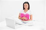 Amused young dark haired model cuddling a pillow and using a laptop in bright bedroom