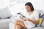 Concentrated young dark haired woman in white clothes reading magazines in a living room