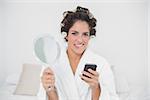 Unsure natural brunette holding mirror and smartphone in bedroom