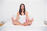 Peaceful young brown haired model in white pajamas practicing yoga in bright bedroom