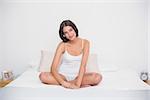 Calm young brown haired model in white pajamas sitting on her bed in bright bedroom