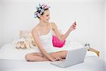 Beautiful natural brown haired woman in hair curlers shopping online with a laptop in bright bedroom