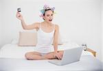 Excited natural brown haired woman in hair curlers shopping online with her laptop in bright bedroom