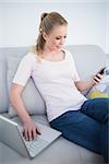 Casual smiling blonde using smartphone and laptop in bright living room