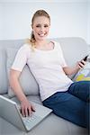 Casual cheerful blonde using smartphone and laptop in bright living room