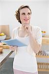 Smiling thinking woman holding her tablet standing in kitchen at home