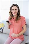 Content calm pregnant woman holding her smartphone sitting on couch smiling at camera