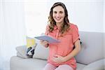 Cheerful pregnant woman holding her tablet sitting on a couch smiling at camera
