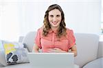 Cheerful pregnant woman using her notebook while sitting on couch and smiling at camera