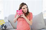 Lovely pregnant woman shaking a piggy bank sitting on couch in the living room