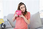 Beautiful pregnant woman shaking a piggy bank sitting on couch in living room