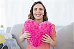 Cheerful pregnant woman holding pink heart pillow sitting on couch looking at camera