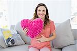 Content pregnant woman holding a pillow sitting on couch in living room smiling at camera