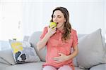 Lovely pregnant woman eating green apple sitting on couch in living room at home
