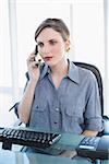 Serious young businesswoman phoning with her smartphone sitting at her desk in the office