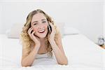 Pretty smiling blonde lying on bed phoning in bright bedroom