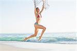 Joyful slender woman jumping in the air holding shawl on the beach