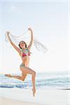 Cheerful slender woman jumping in the air holding shawl on the beach