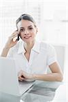 Smiling young businesswoman phoning with her smartphone looking at camera sitting at her desk