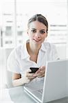 Lovely businesswoman holding her smartphone smiling softly at camera sitting at her desk