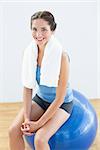 Portrait of a smiling young woman with towel around neck sitting on exercise ball